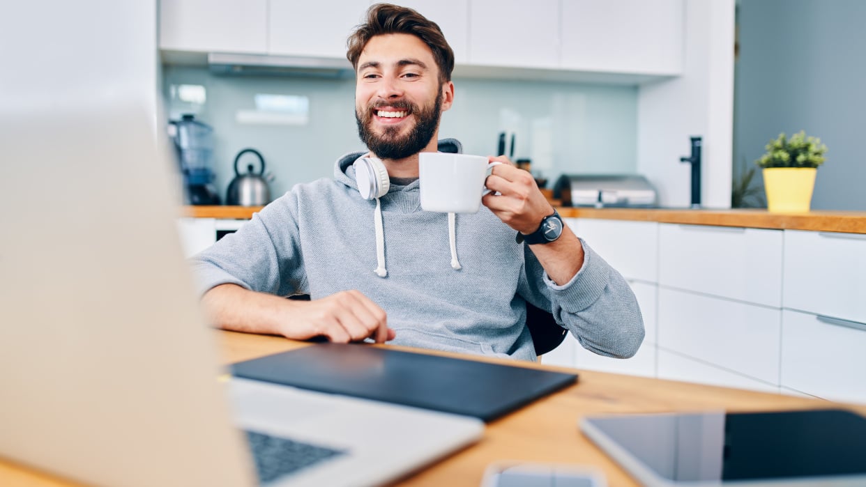 Mann hält Kaffeetasse und lächelt zufrieden auf den Bildschirm seines Laptops